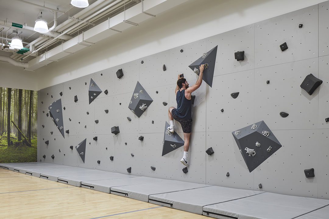 A man in athletic wear climbs an indoor bouldering wall in a gym, using large geometric holds. Safety mats cover the floor, and a forest-themed mural is visible in the background.