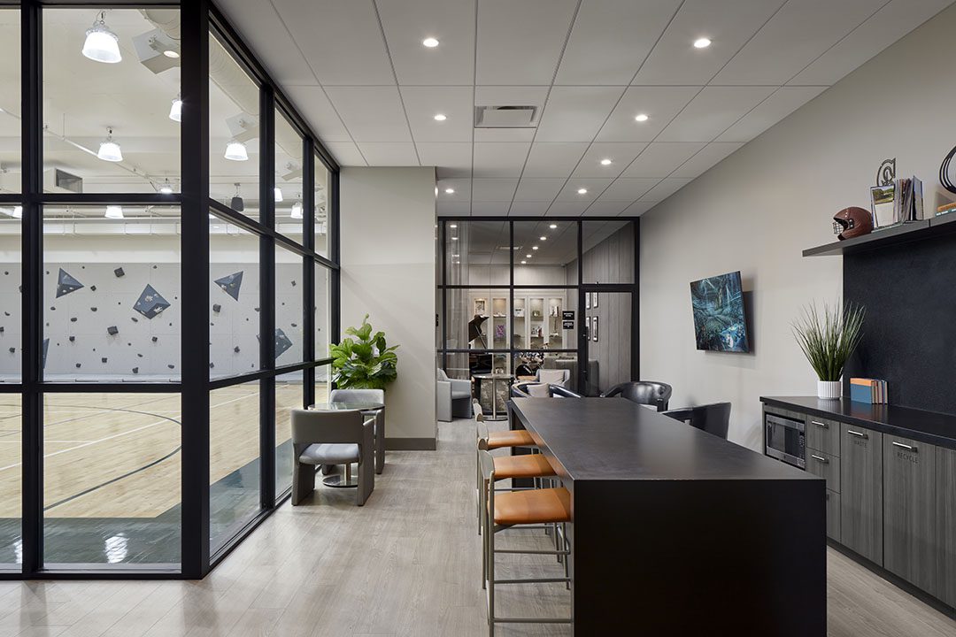 Modern office lounge with a high table, orange chairs, and cabinets, adjacent to a gym with a basketball court and rock climbing wall, separated by large glass windows. Decor includes plants and sports memorabilia.