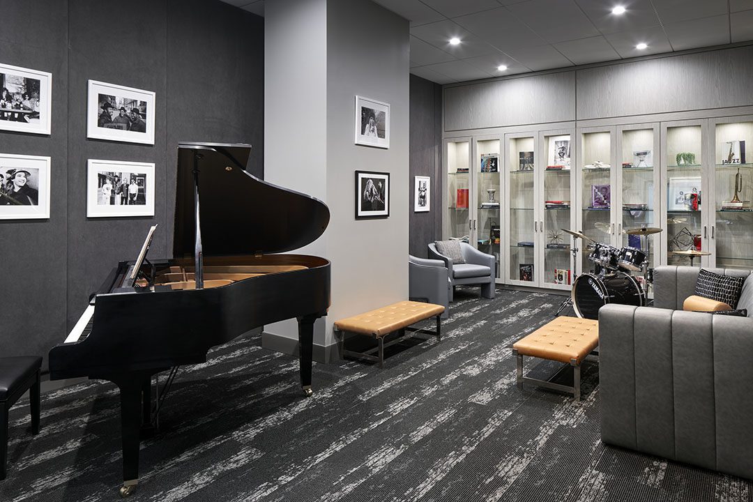 Modern music room with a grand piano, drum set, gray walls decorated with framed photos, glass display cabinet with memorabilia, a gray chair, and cushioned benches on a patterned carpet.