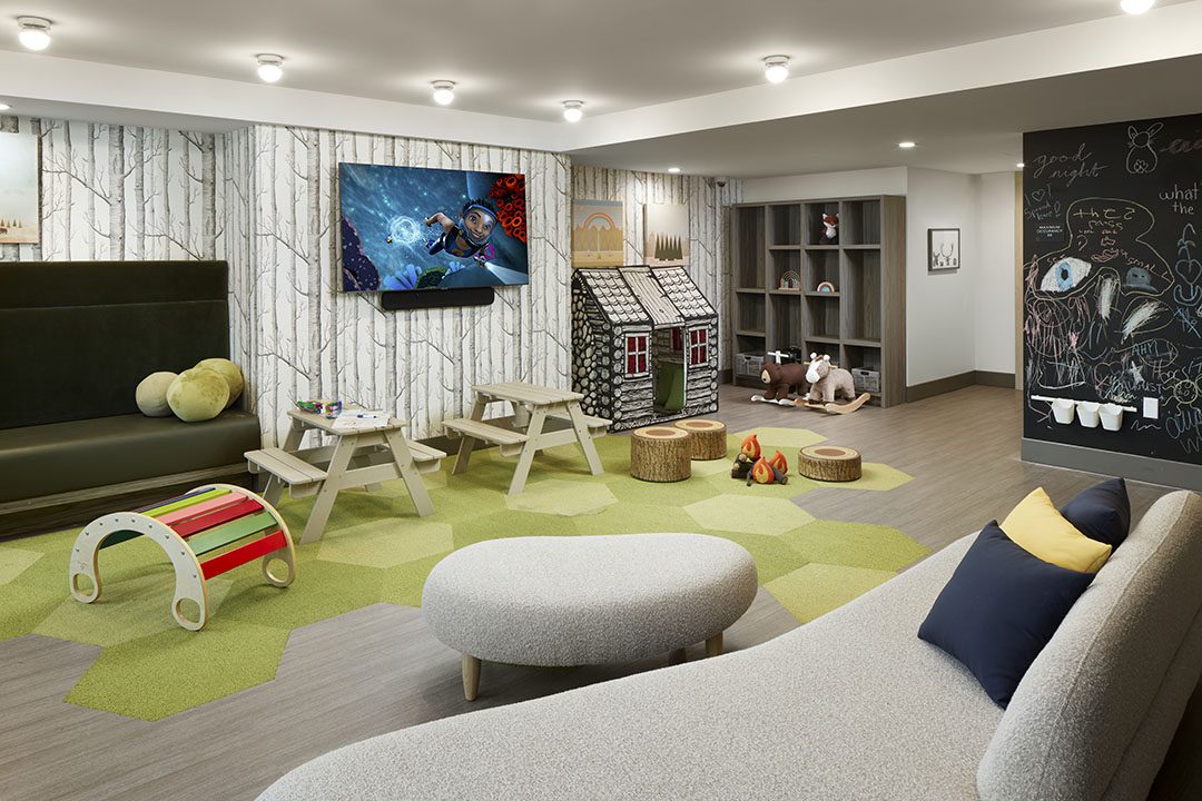 A modern childrens playroom with toys, a TV on the wall, playhouses, child-sized tables, a chalkboard, and cozy seating, decorated with playful accents and green geometric carpet tiles.