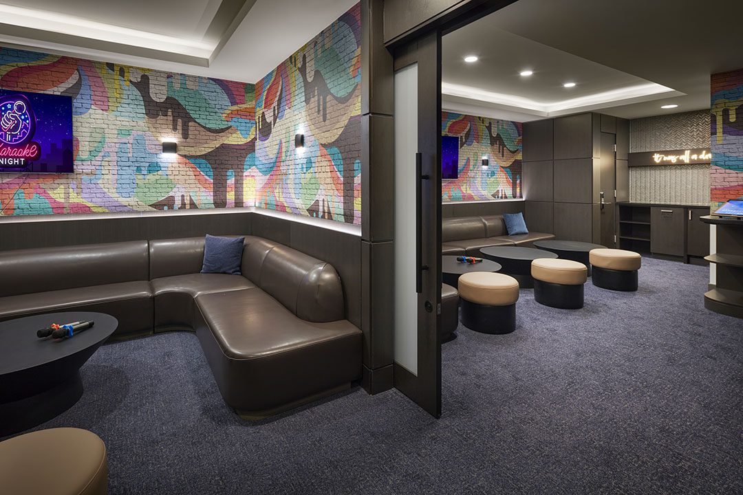 Modern karaoke lounge with a brown L-shaped sofa, colorful graffiti-style wall art, small round tables, and stools. Neon signs and dim lighting create a cozy atmosphere. A private room with a bar area is visible in the background.