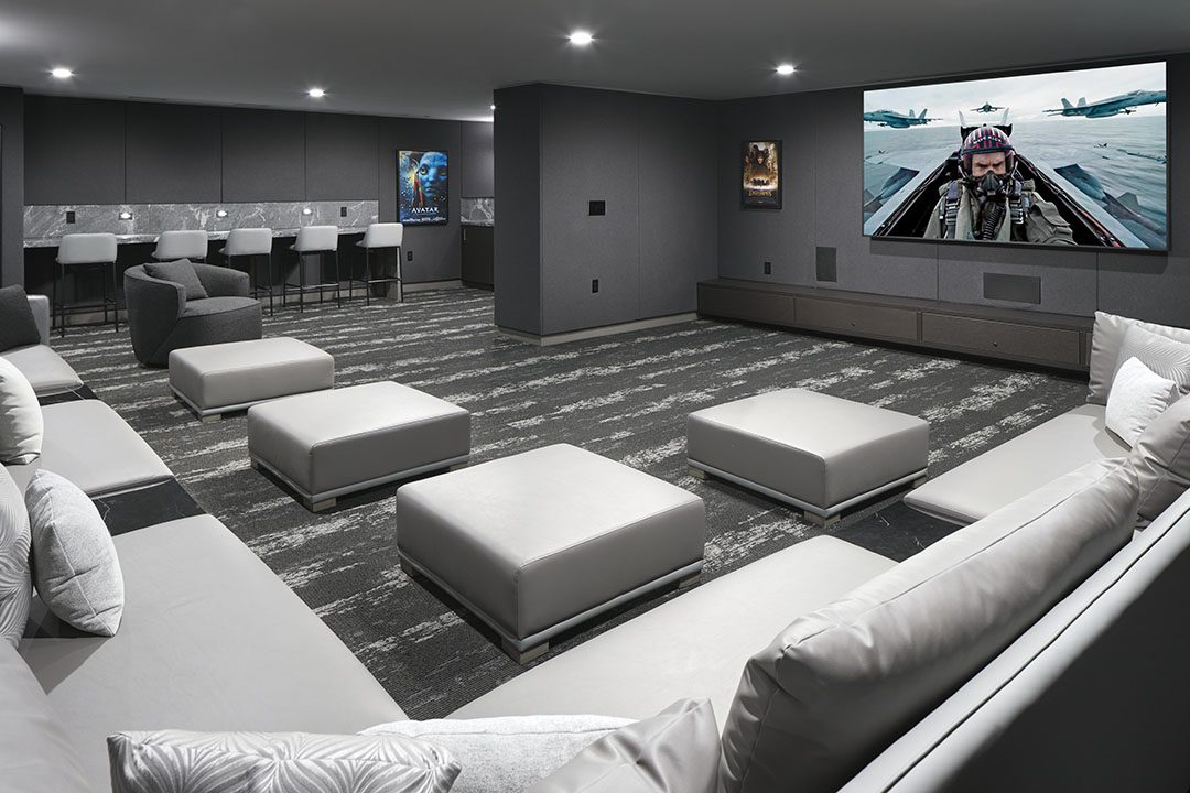 Modern home theater room with gray couches, ottomans, a large screen showing a fighter jet scene, wall posters, bar seating area, and minimalist gray decor.