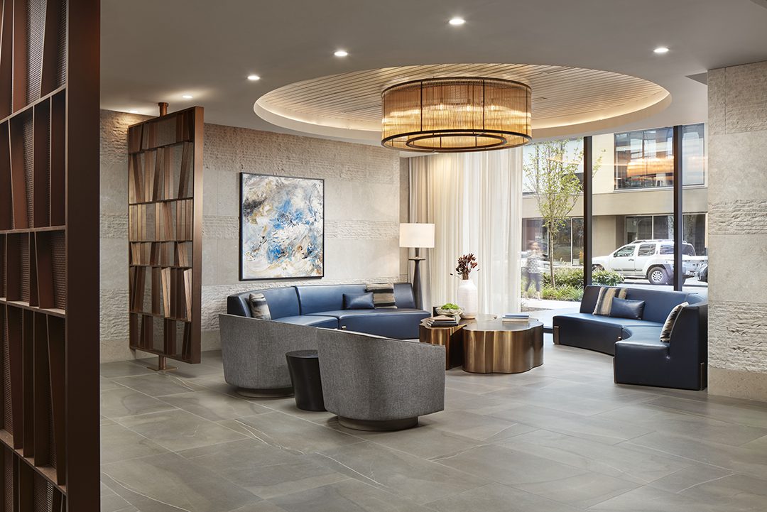 Modern lounge area with blue and gray seating, round coffee tables, large abstract wall art, decorative light fixture, and floor-to-ceiling windows showing cars and trees outside. Contemporary decor and sleek, neutral tones throughout.