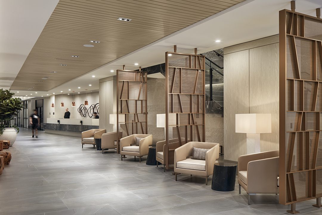 Modern hotel lobby with beige armchairs and wooden room dividers, marble floor, and contemporary decor. Soft lighting and minimalist design create a welcoming atmosphere. A reception desk is visible in the background.