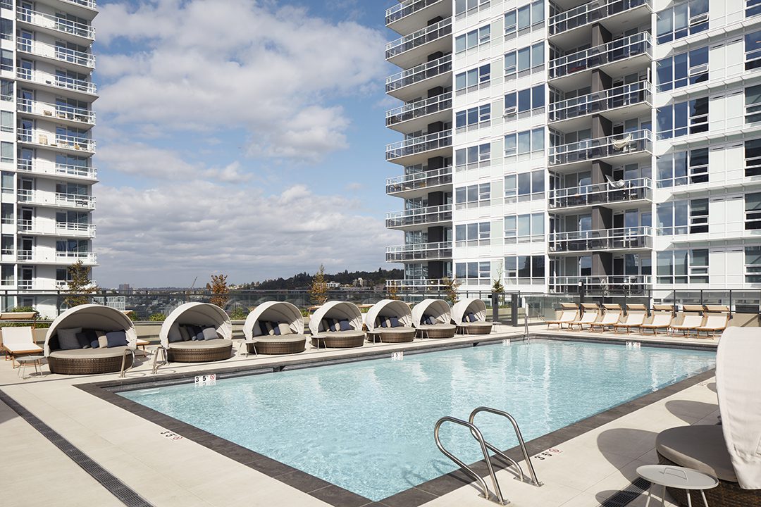 A modern outdoor swimming pool surrounded by lounge chairs and cabana beds, set between tall white apartment buildings under a partly cloudy sky.