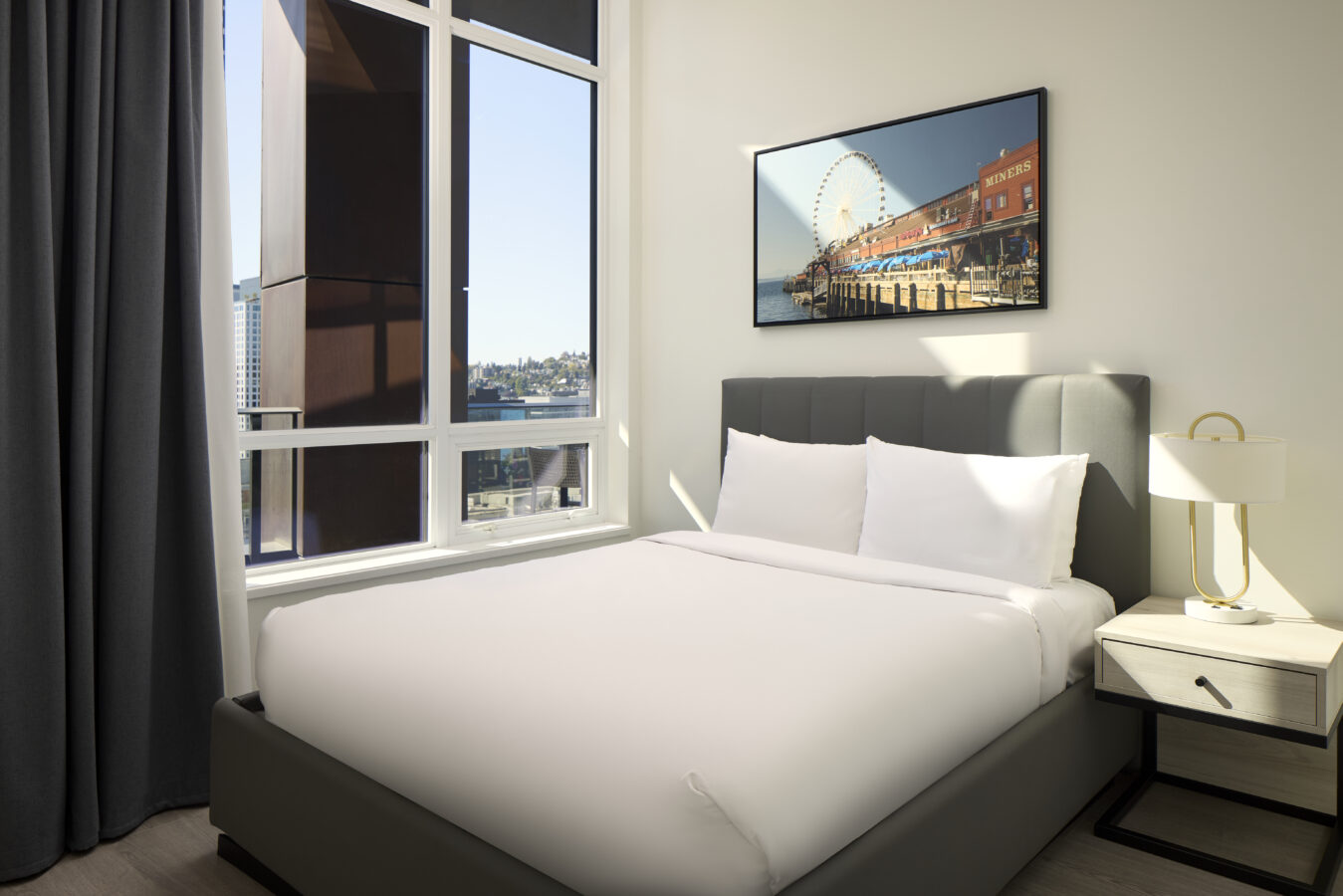 A modern bedroom with a large window, a neatly made bed with white linens, a nightstand with a lamp, dark curtains, and a framed photo of a Ferris wheel and waterfront scene on the wall.