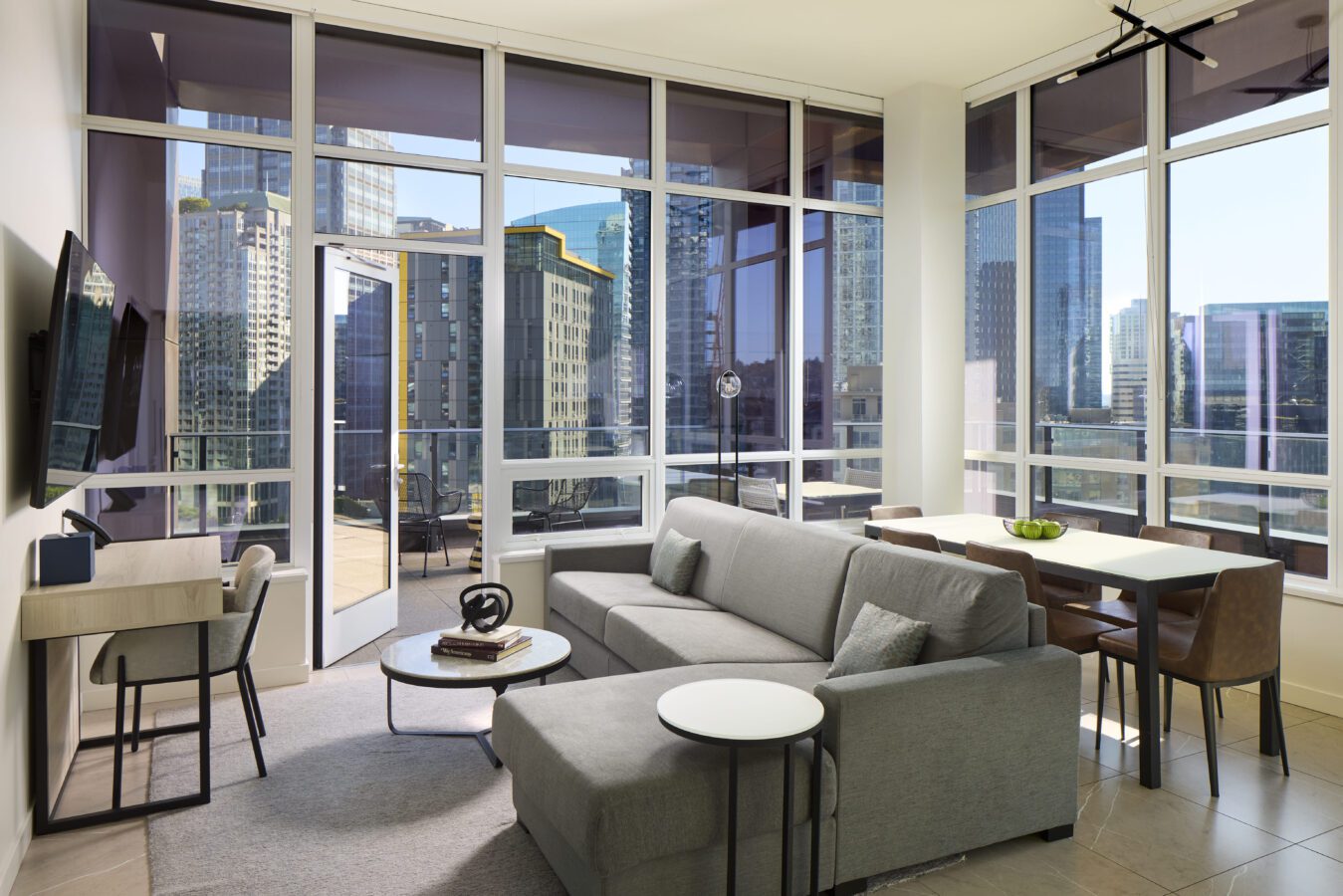 Modern living room with large glass windows overlooking a city skyline, featuring a gray sectional sofa, TV, desk, dining table with chairs, and abundant natural light streaming in.