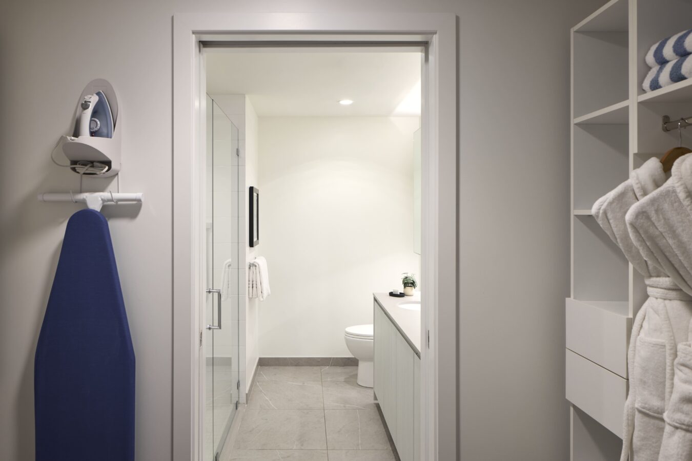 A modern bathroom viewed through a doorway, featuring a toilet, sink with a countertop, mirror, and a glass shower. In the foreground, an ironing board and robe hang on the wall, with shelves holding towels to the right.