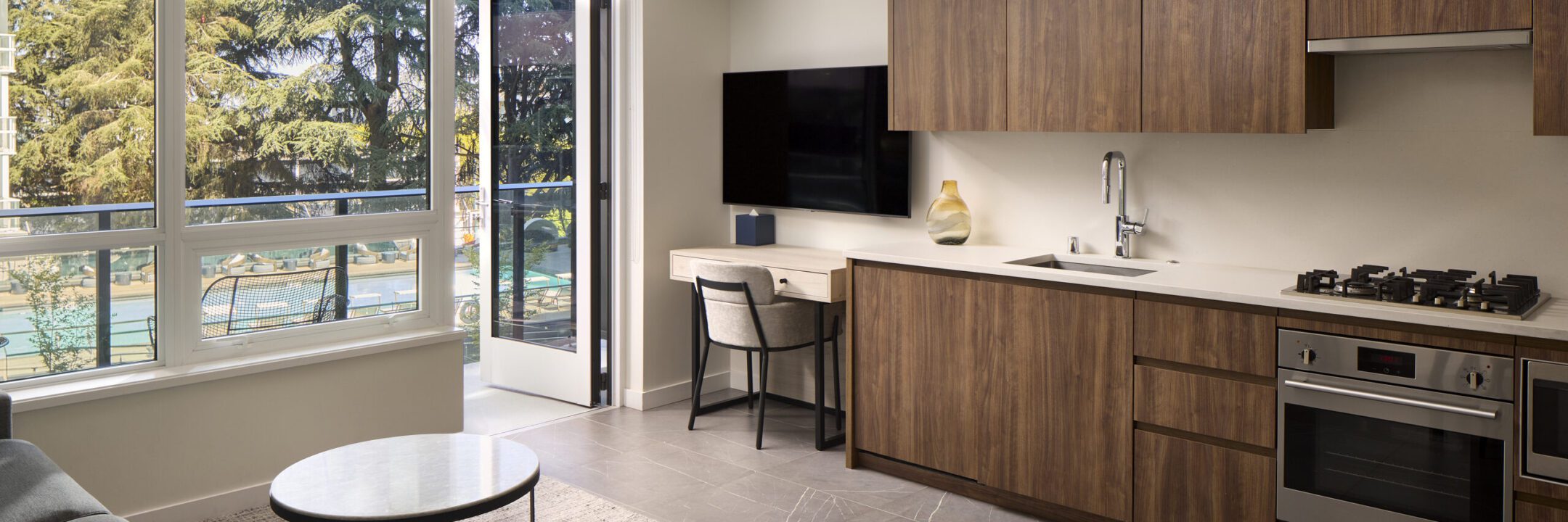 Modern kitchen and workspace with wooden cabinets, built-in oven, desk, chair, wall-mounted TV, and large window showing an outdoor view with trees and a sports court.