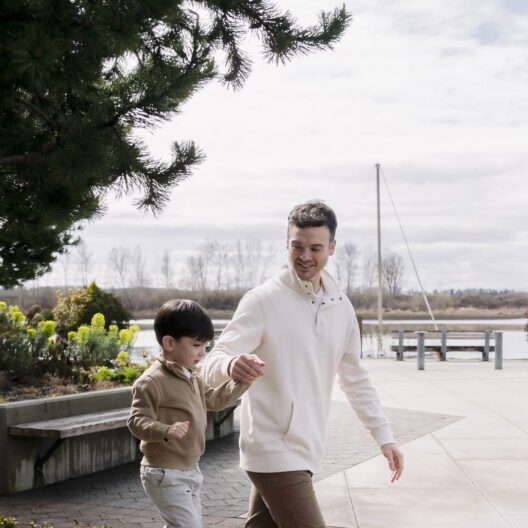 A man and a young boy walk hand in hand on a paved path near a park with trees and water in the background. Both are smiling and dressed in casual, light-colored clothing.