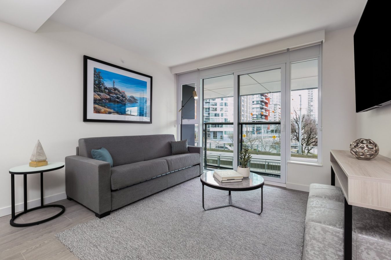 Modern living room with a gray sofa, round coffee table, wall art, and large windows overlooking city buildings. Light decor with a small side table and a gray rug on the floor.