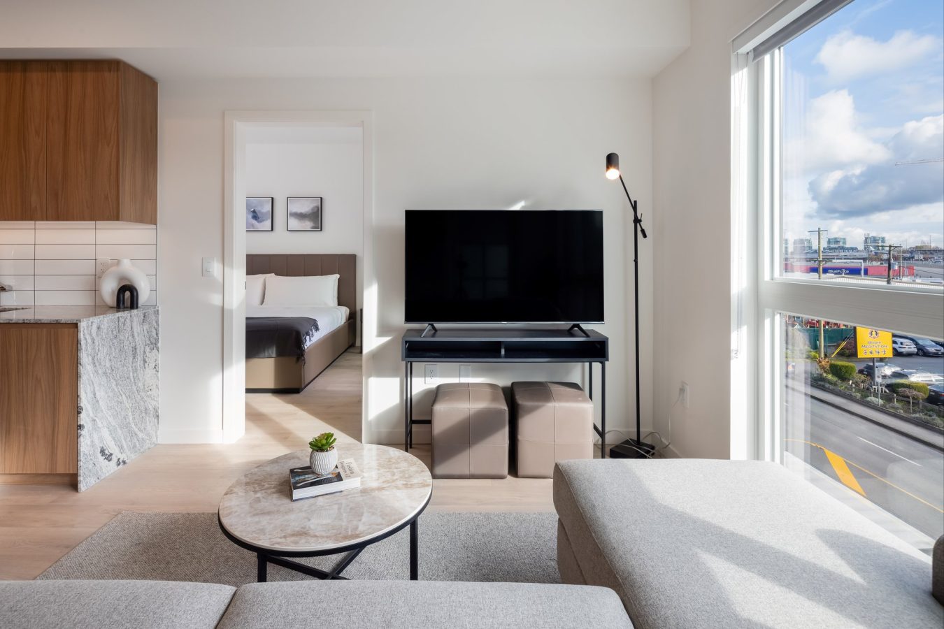 Bright, modern living room with a gray sectional, round marble coffee table, TV on a stand, floor lamp, large window with city view, kitchen area with wood cabinets, and a bedroom visible through an open door.