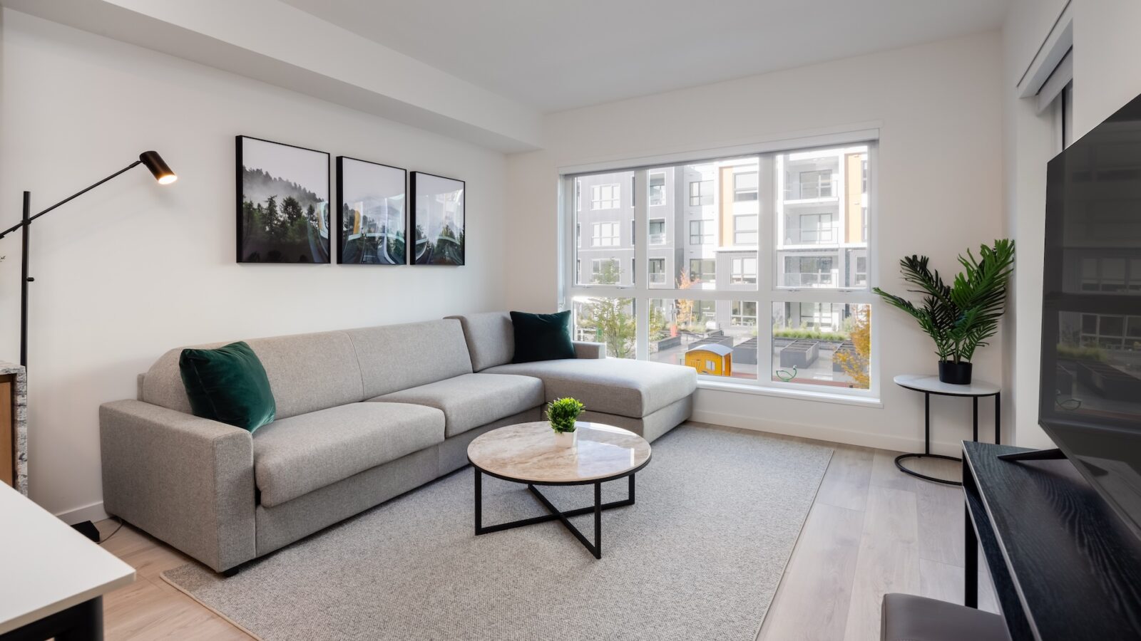 A modern, bright living room with a gray sofa, green accent pillows, a round marble coffee table, wall art, a floor lamp, a potted plant, and large windows showing an apartment building outside.