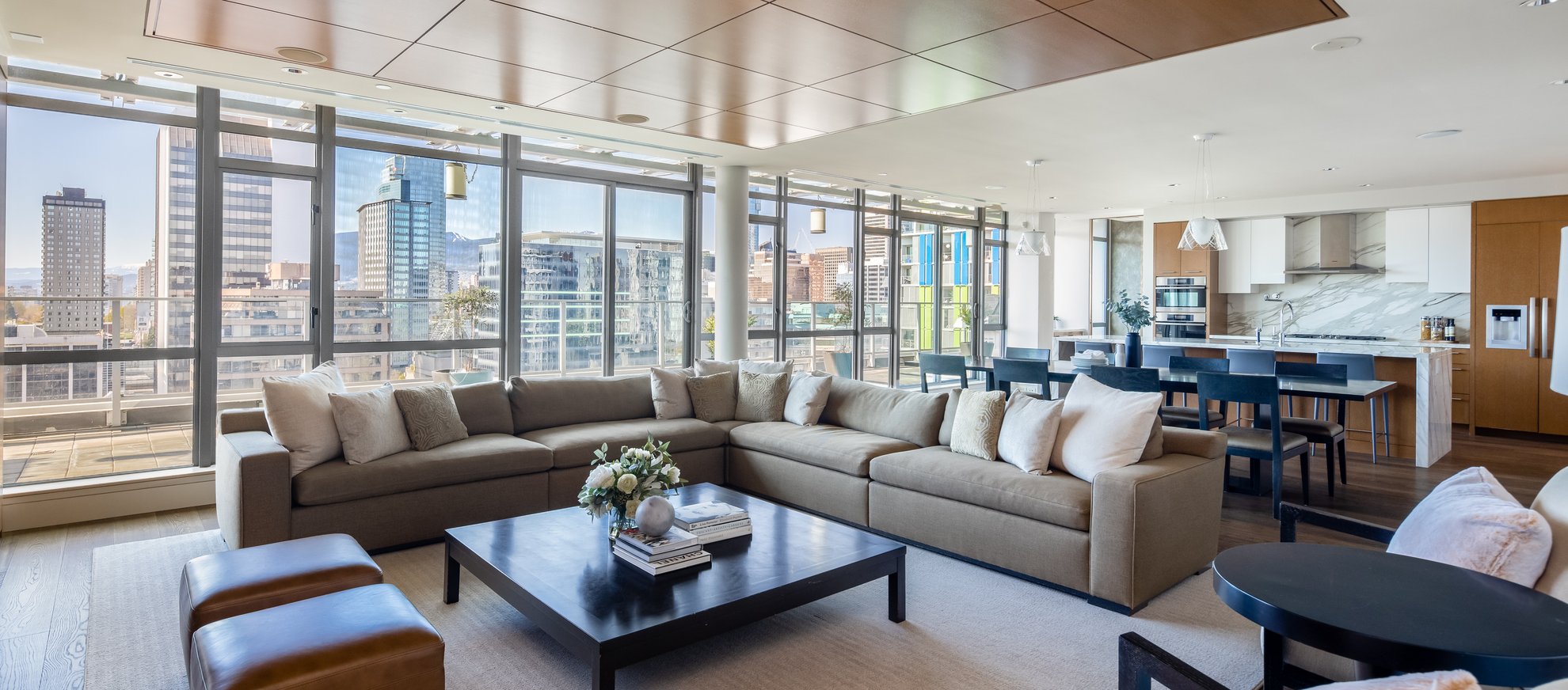 Modern, spacious living room and kitchen with large windows offering city views, neutral-toned sofas, a coffee table with books and flowers, a dining table with chairs, and sleek kitchen appliances in an open-plan layout.