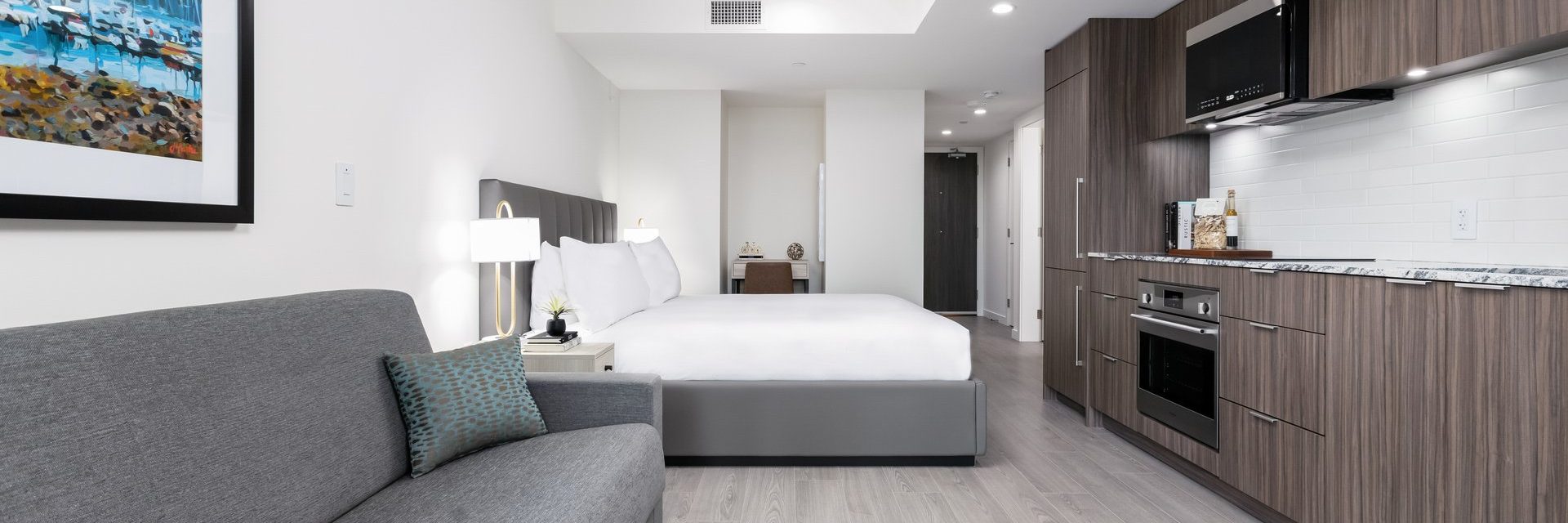 Modern studio apartment with a bed, gray sofa, nightstand, wall art, and a kitchenette featuring wood cabinets, a microwave, and marble countertops. The space is bright with white walls and contemporary decor.