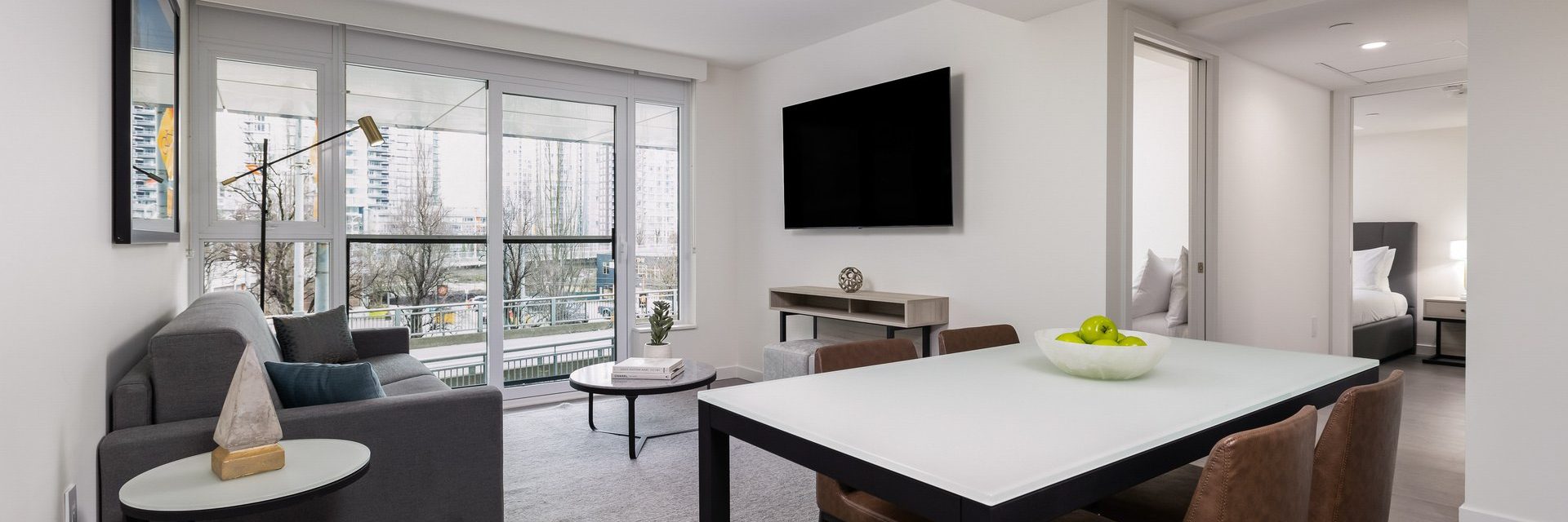 Modern apartment living room with large windows, a gray sofa, wall-mounted TV, dining table with a bowl of green apples, and a view of tall buildings outside. A bedroom is visible through an open doorway.