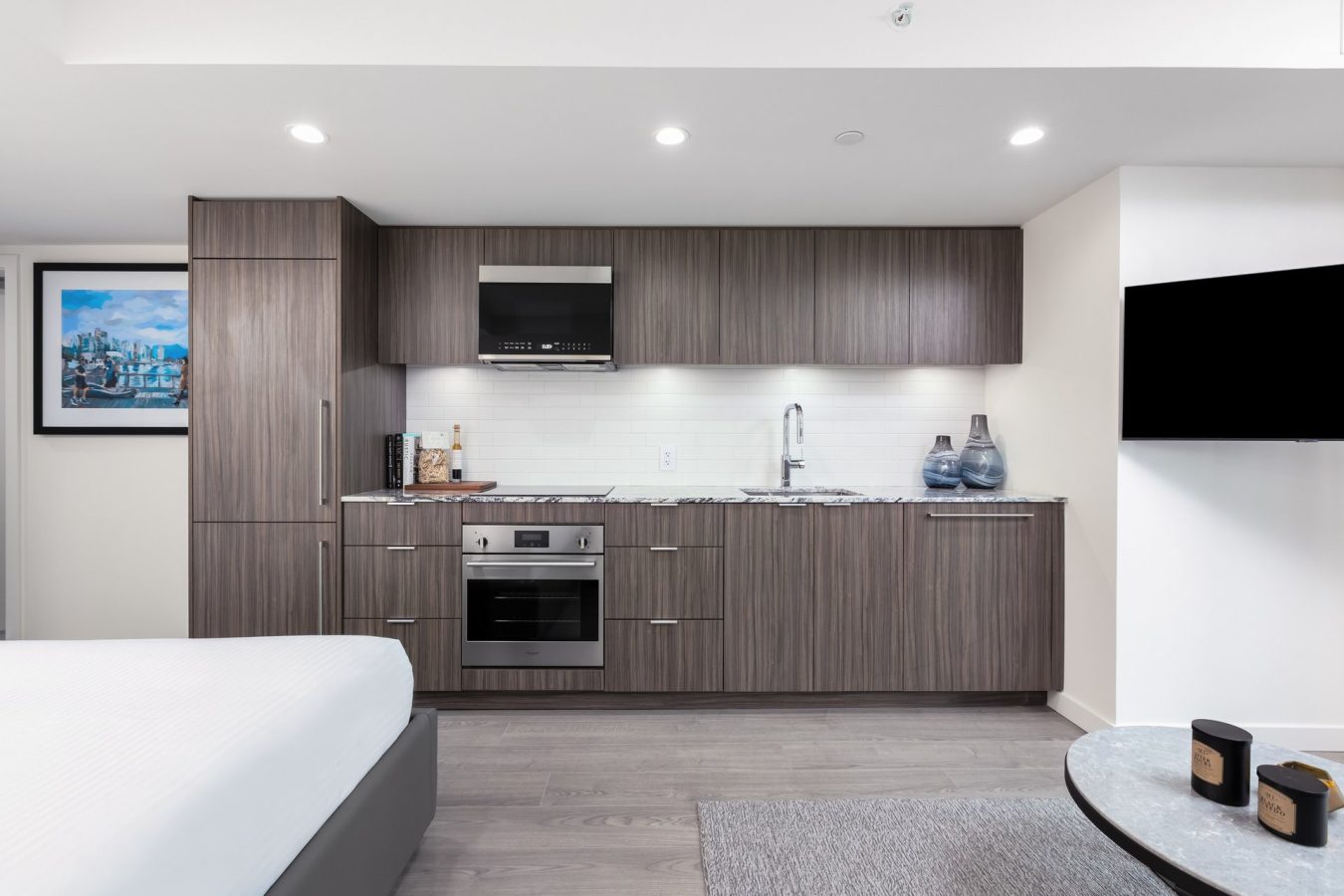 Modern studio apartment kitchen with wood cabinets, built-in oven, microwave, sink, vases, and wall-mounted TV; part of a bed and a coffee table with candles are also visible in the open-plan space.