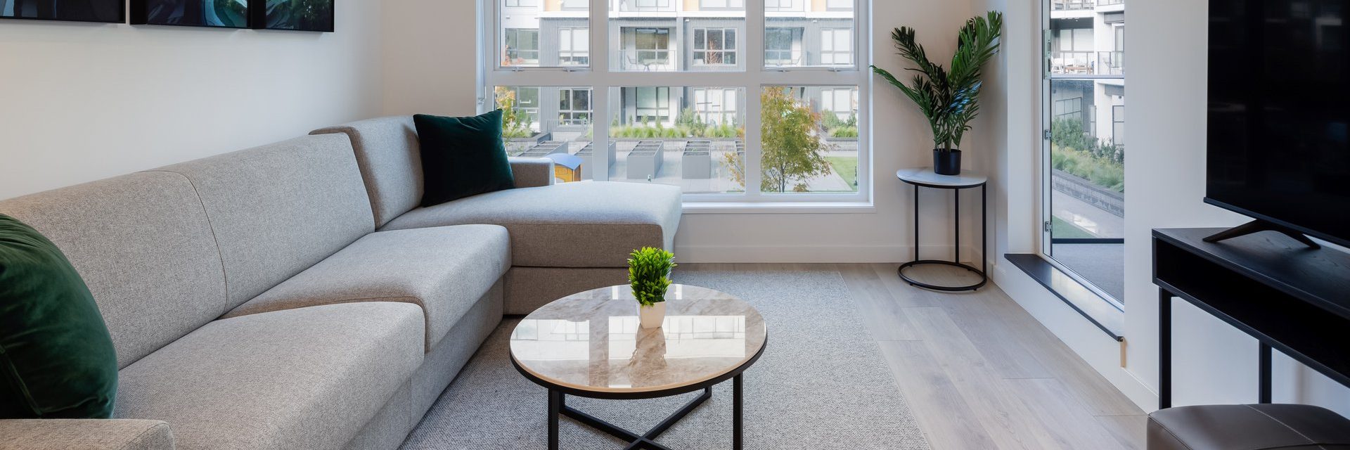 A modern living room with a light gray sectional sofa, a round marble coffee table with a small plant, a potted plant on a side table, and large windows letting in natural light.