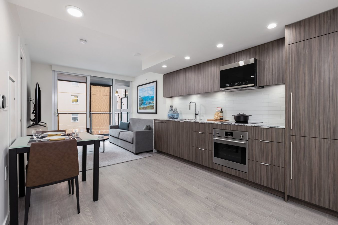Modern studio apartment with a sleek kitchen, gray cabinets, built-in appliances, a small dining table set for two, a gray sofa, wall art, and large windows letting in natural light.