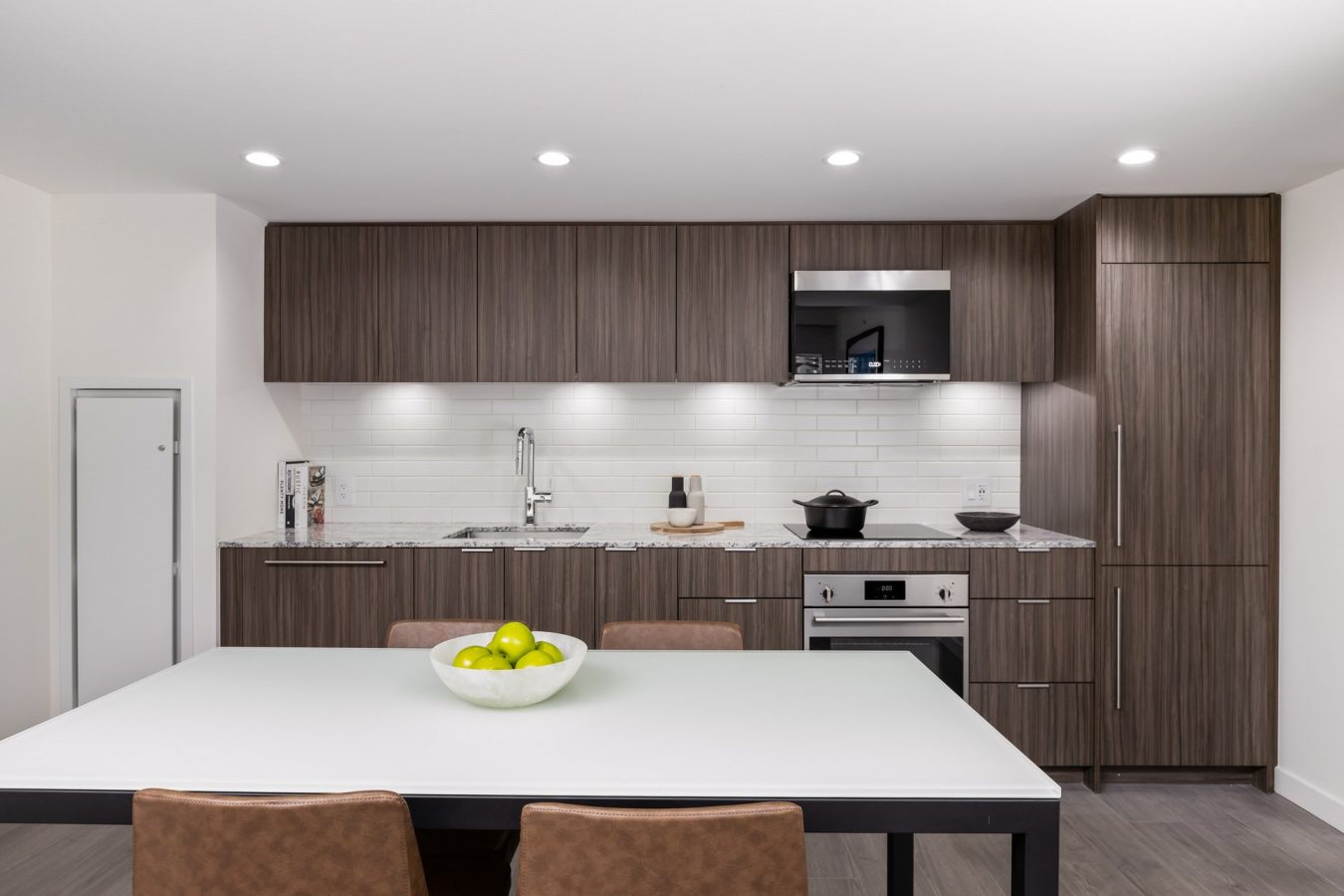 A modern kitchen with wood cabinets, white countertops, a built-in oven, stove, sink, and microwave. On the dining table in front, there are four brown chairs and a bowl of green apples.