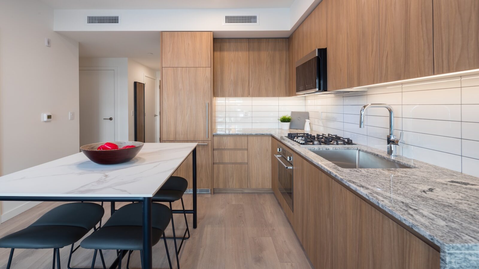 Modern kitchen with wood cabinets, marble countertops, stainless steel appliances, gas stove, and a white subway tile backsplash. A marble dining table with four black chairs and a bowl of red apples is in the foreground.