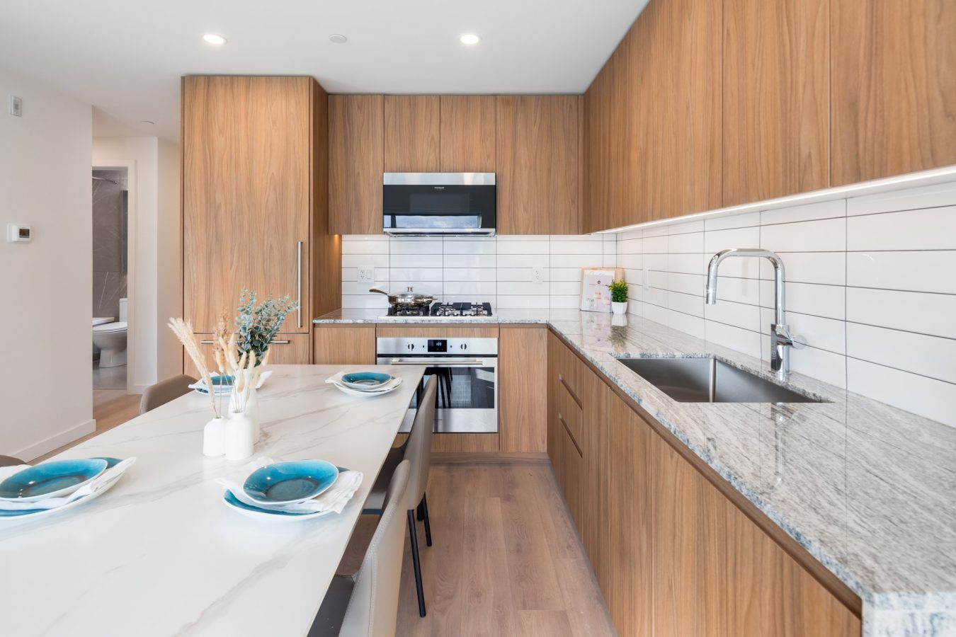 Modern kitchen with wooden cabinets, marble countertops, a built-in oven and microwave, gas stove, stainless steel sink, and a dining area set with blue plates and white vases on a white table.