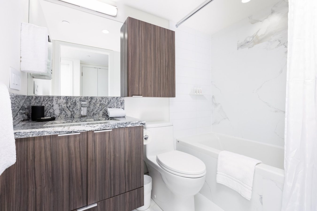 Modern bathroom with wood-tone cabinets, marble countertop, white toilet, bathtub with marble-patterned wall, and a white shower curtain. A mirror and towel are also visible, creating a clean and bright look.