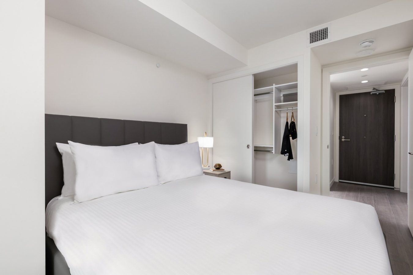 A modern, minimal bedroom with a large bed, white bedding, a padded headboard, a lamp on a bedside table, an open closet with hangers, and a hallway leading to a closed door.