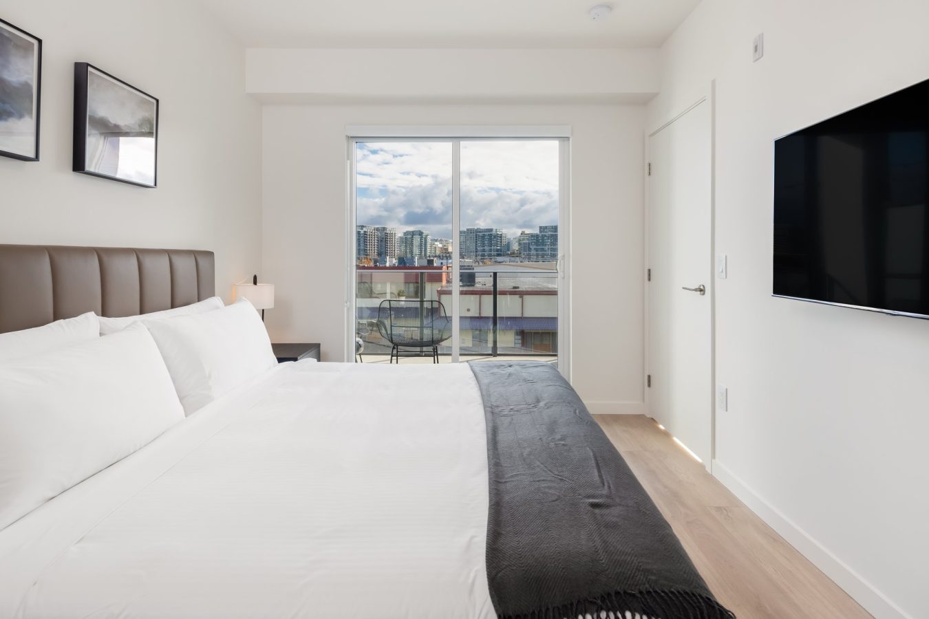 A modern, minimal bedroom with a large bed, white linens, a dark throw blanket, wall-mounted TV, and a sliding glass door leading to a balcony with city buildings visible outside.