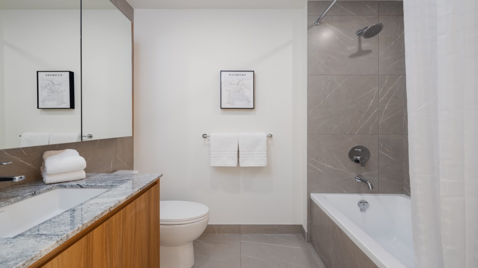 A modern bathroom with a marble countertop, wooden vanity, wall mirror, toilet, towel rack with white towels, bathtub with shower, gray tile walls, and framed artwork on a white wall.