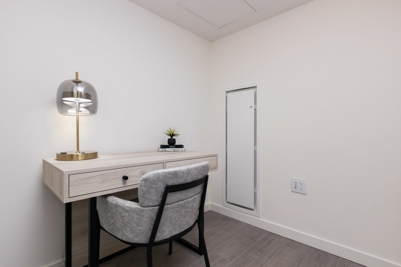 A small, minimalist home office features a light wood desk with a gray chair, a modern lamp, a small potted plant, and stacked books, set against plain white walls and gray wood flooring.