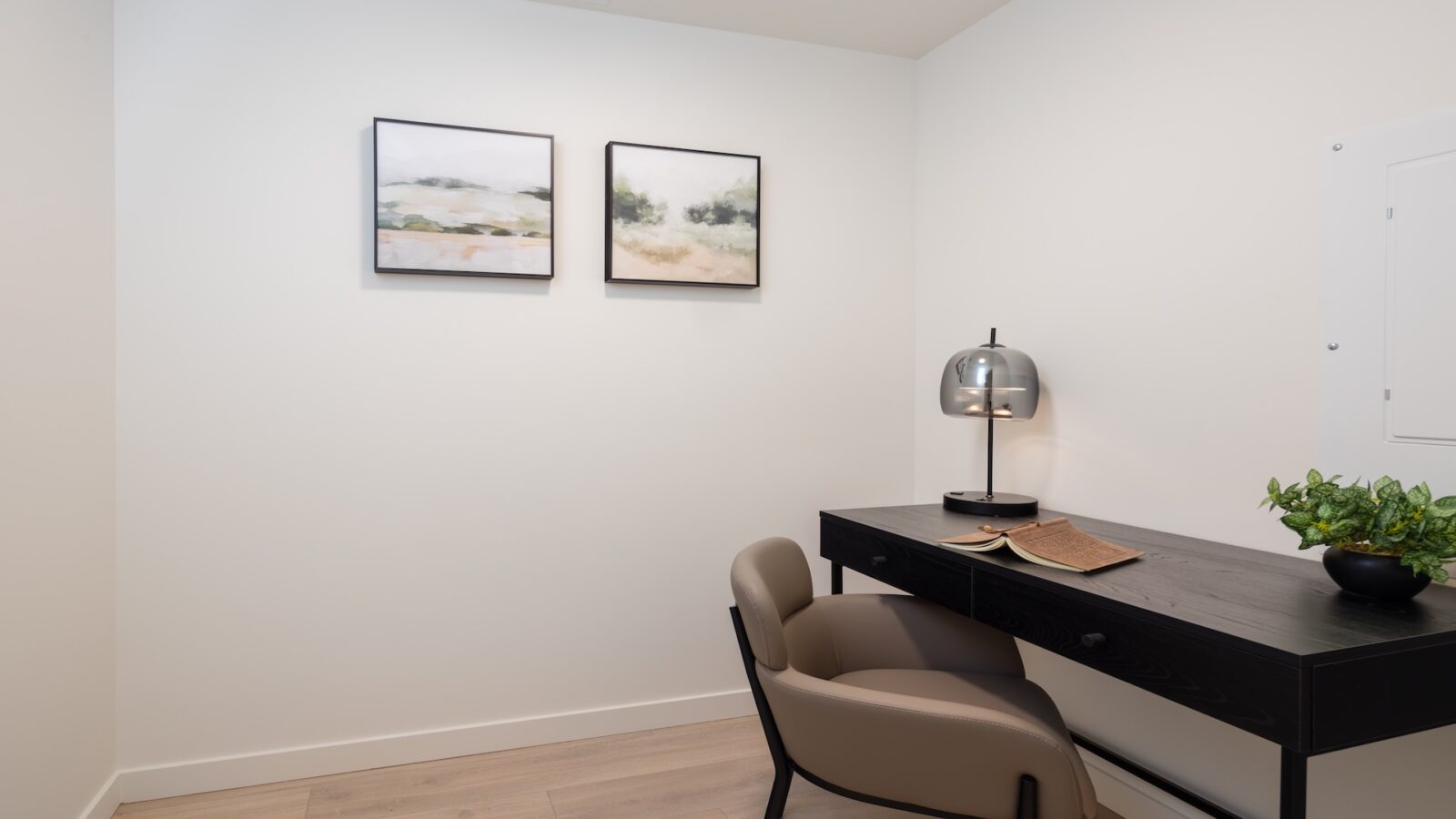 A minimalist home office with a black desk, beige chair, lamp, open book, potted plant, and two abstract landscape paintings on the white wall. Light wood flooring completes the clean, modern look.