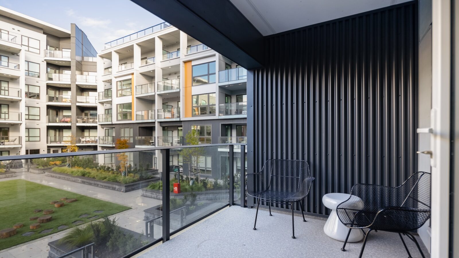 Modern apartment balcony with two black chairs and a small white table, overlooking a courtyard with green grass and garden beds. Glass railing and contemporary multi-story buildings are visible in the background.