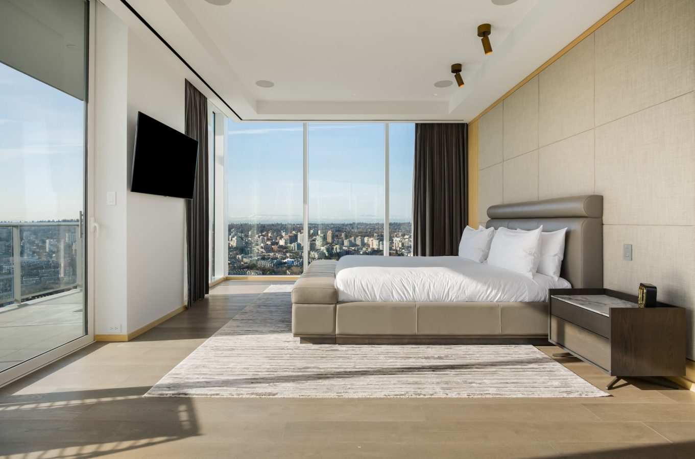 Modern bedroom with a large bed, white bedding, floor-to-ceiling windows with city views, a wall-mounted TV, and minimalist furniture on a neutral-toned rug. Natural light fills the spacious, elegant room.