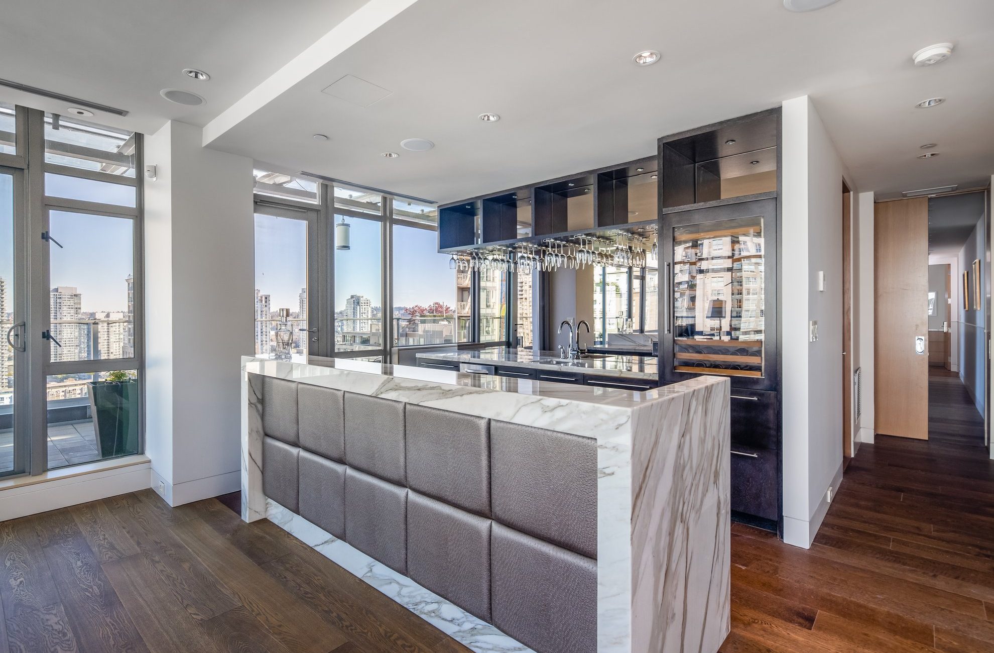 Modern apartment interior with a marble breakfast bar, padded front panel, wall-mounted glass racks, floor-to-ceiling windows, and city skyline views. Hardwood floors and sleek cabinetry complete the contemporary look.