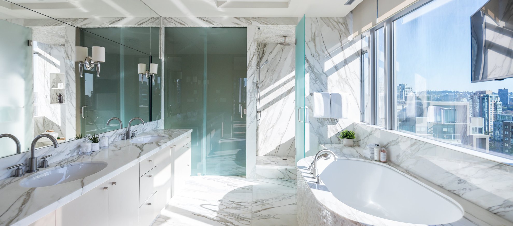 Modern, luxurious bathroom with marble surfaces, double sinks, a large bathtub by a window with city views, glass shower enclosure, and bright natural light streaming in.