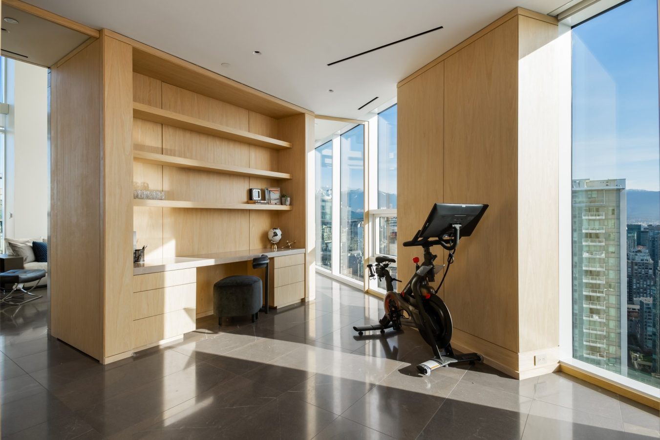 Modern home office and workout area with built-in wooden shelves and desk, black chair, and exercise bike beside floor-to-ceiling windows overlooking a cityscape with tall buildings and mountains.