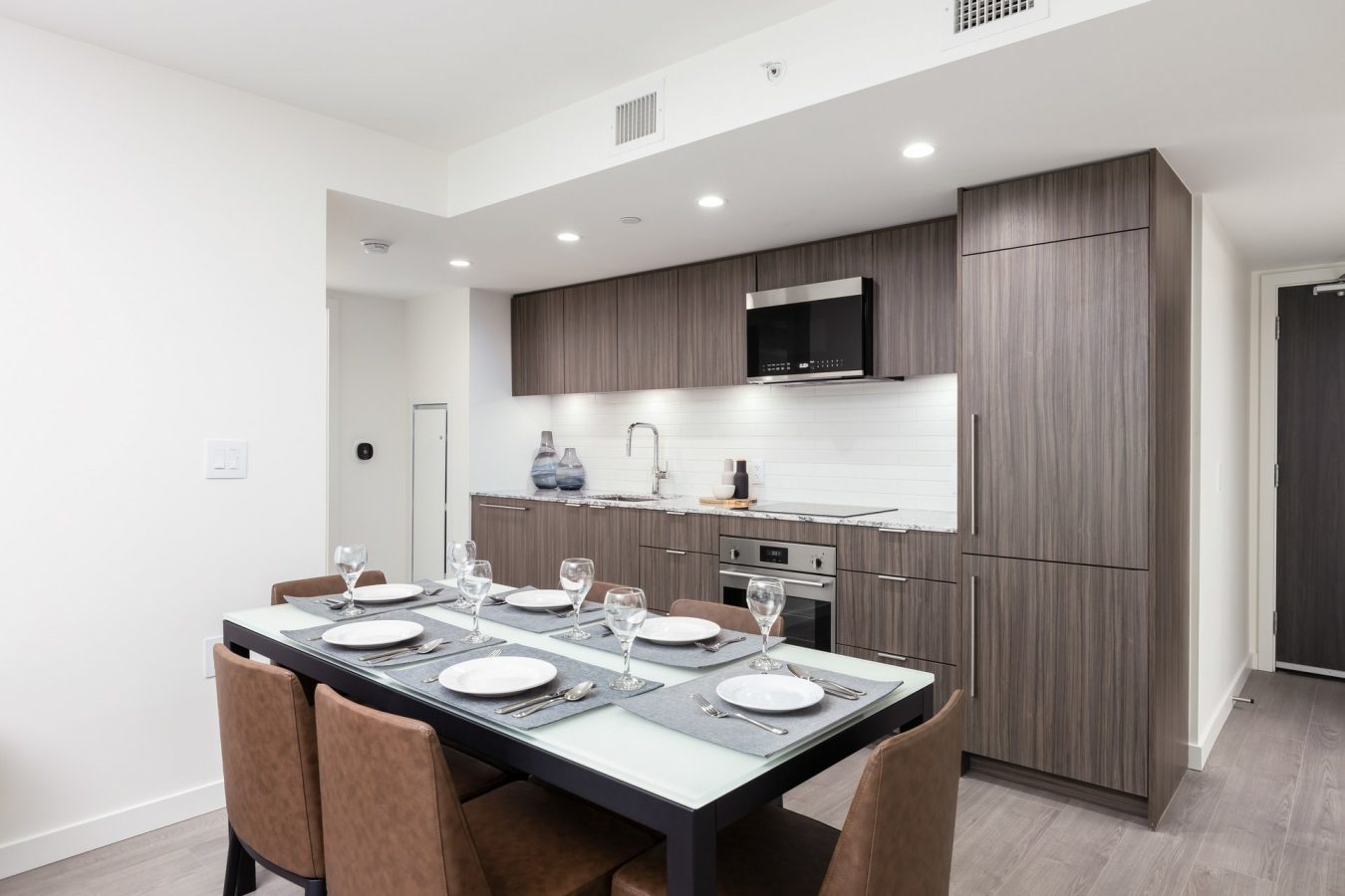 A modern kitchen with wood cabinets, built-in appliances, and a white tile backsplash. In the foreground, a dining table is set with plates, glasses, and cutlery for six people. The space has a clean, minimalist design.