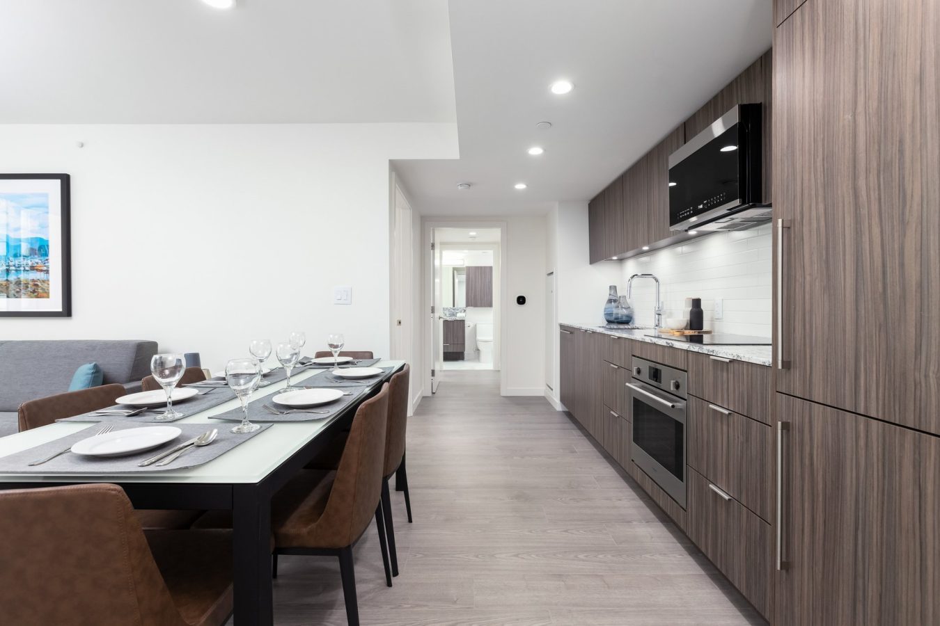 Modern kitchen and dining area with wooden cabinets, a built-in oven, a set dining table with brown chairs, and bright overhead lighting, leading to a hallway and a bathroom in the background.