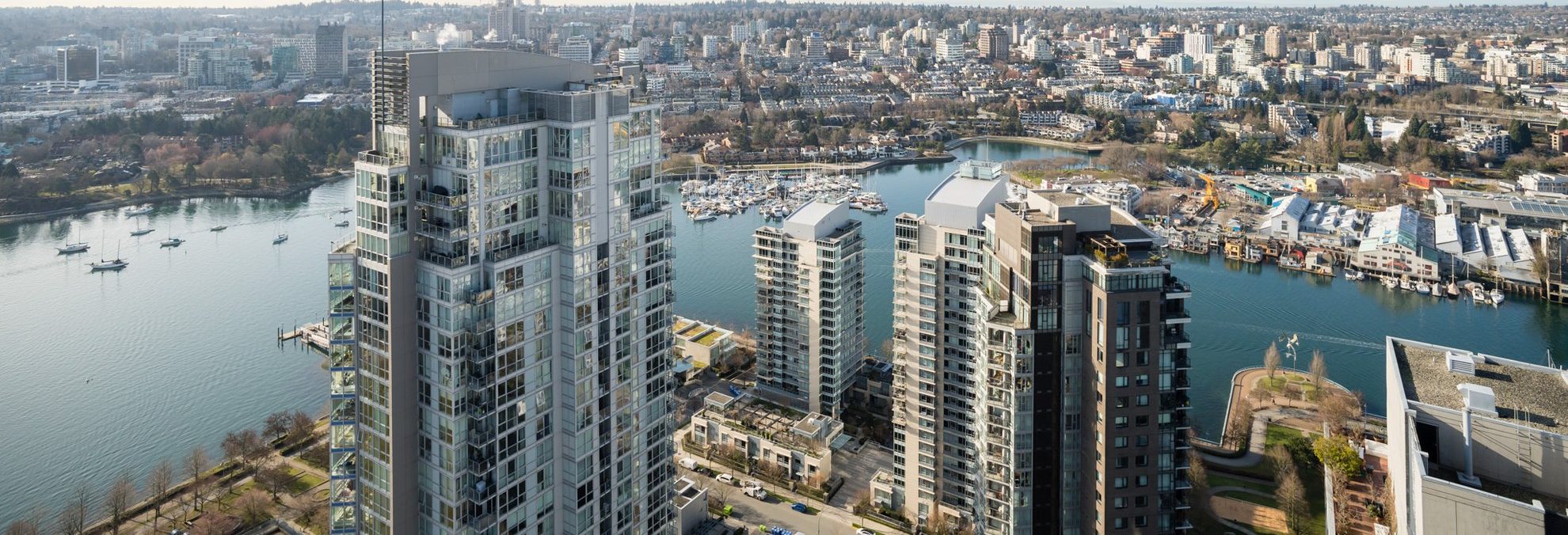 Aerial view of high-rise residential buildings by a waterfront with boats, a marina, and a cityscape in the background under clear skies.