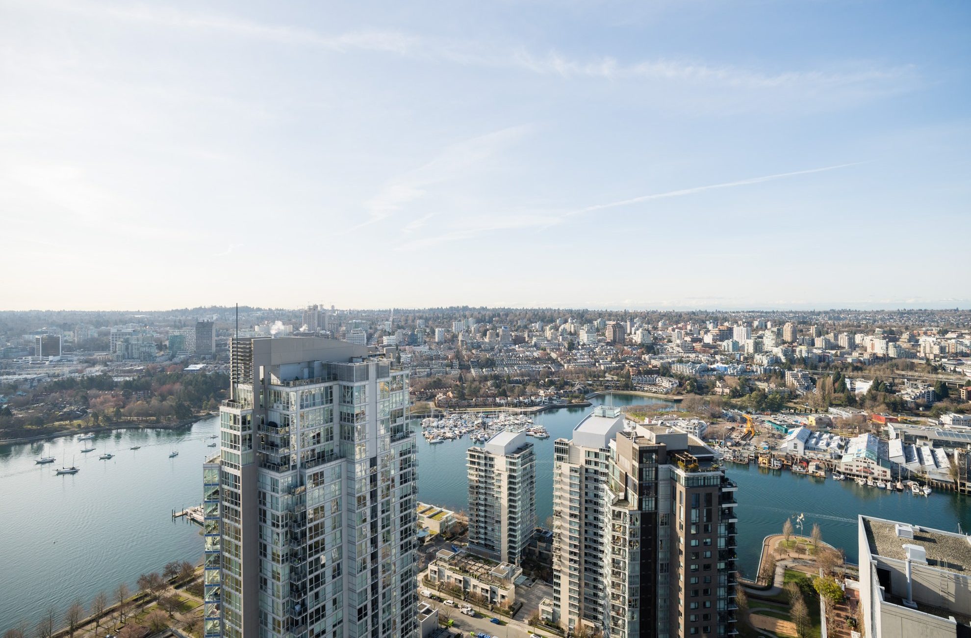 Aerial view of a cityscape featuring modern high-rise buildings near a river or marina, with boats docked and a sprawling urban area in the background under a clear sky.