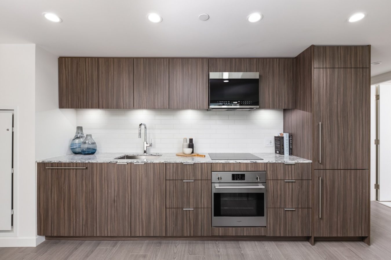 Modern kitchen with flat-panel wood cabinets, marble countertop, built-in oven, electric cooktop, microwave, sink, and under-cabinet lighting. Decorative vases and minimal accessories are on the counter.