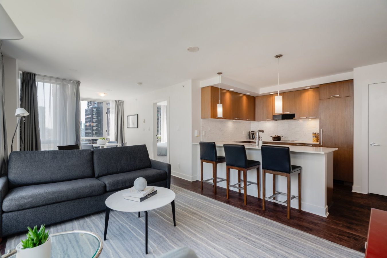 Modern open-concept living space featuring a dark gray sofa, round coffee table, kitchen with island seating, wooden cabinets, and pendant lights. Large windows provide natural light and cityscape views.