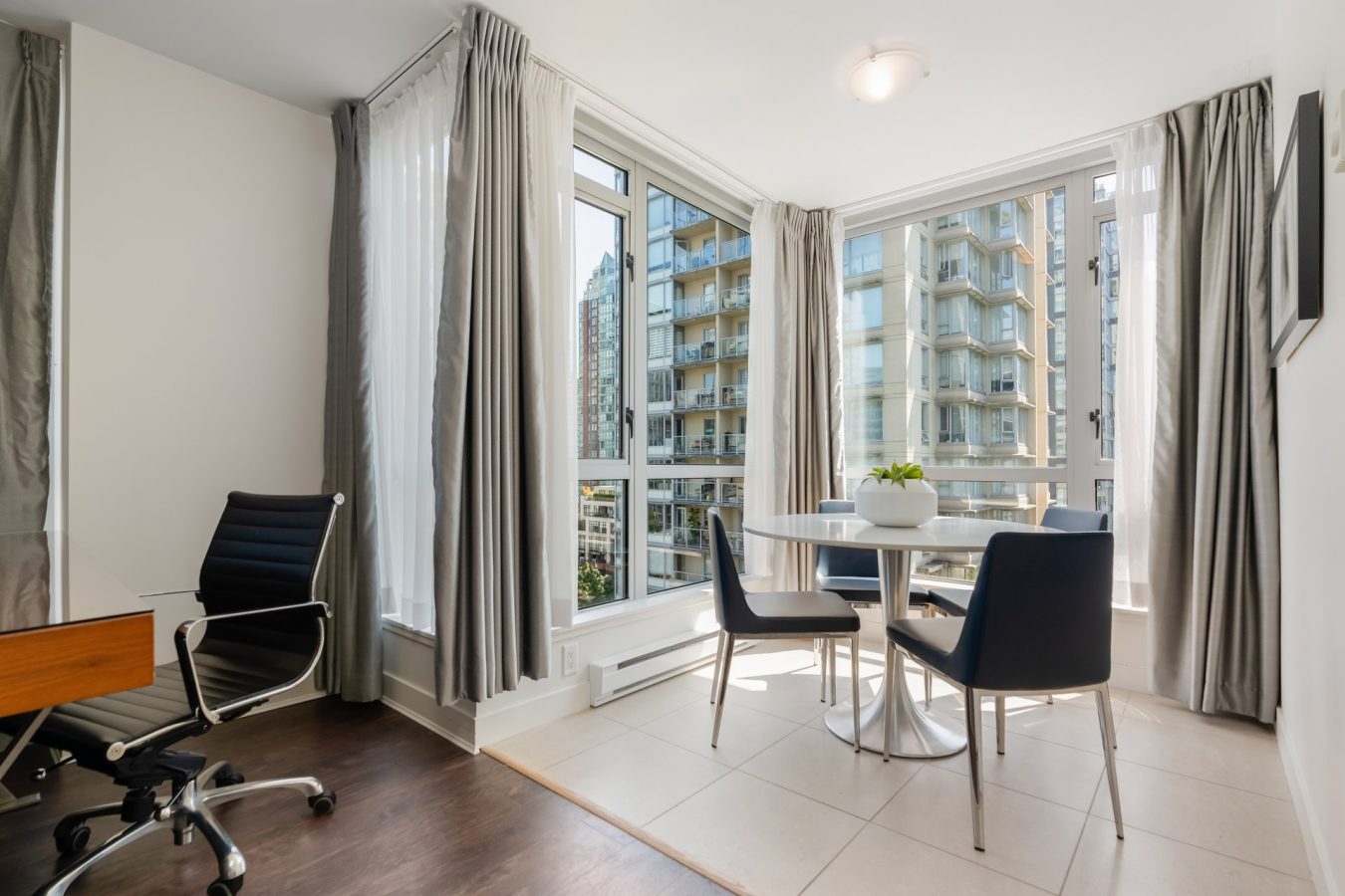 A modern, bright office corner with large windows, a round white table with three black chairs, a potted plant, and a nearby desk with a black office chair. City buildings are visible outside.