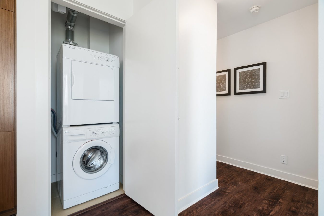 A small laundry closet with a stacked white washer and dryer set, partially enclosed by white walls, next to a hallway with dark wood flooring and two framed pictures on the wall.