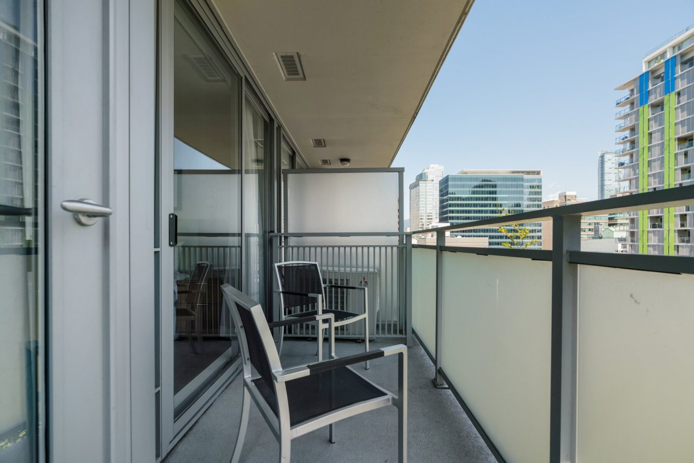 A modern apartment balcony with two black and white chairs, a small table, glass railings, and views of nearby city buildings under a clear blue sky.