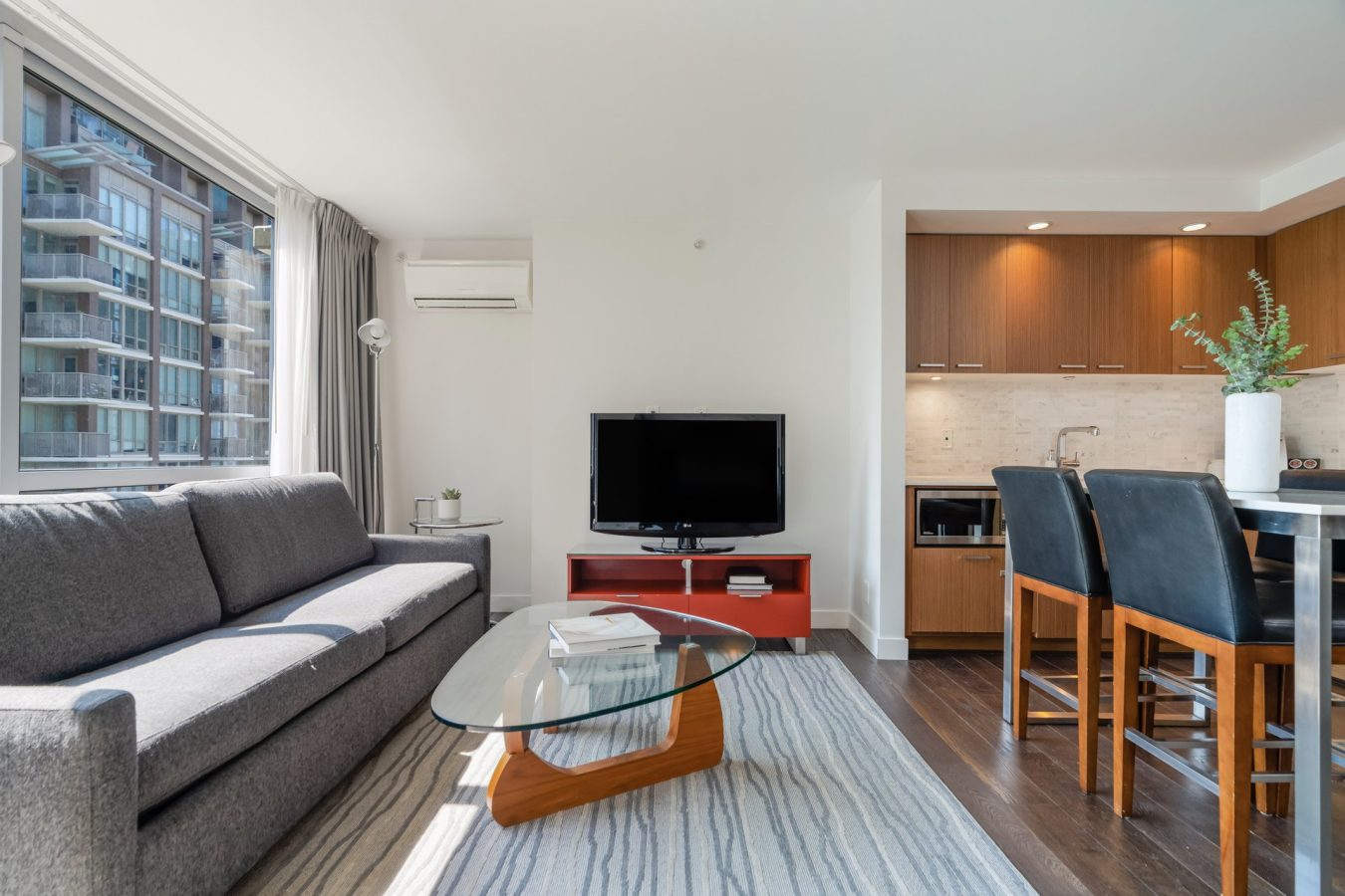 Modern living room with a gray sofa, glass coffee table, TV on a red stand, and an open kitchen with wooden cabinets, bar stools, and a vase with greenery. Large window allows natural light into the space.
