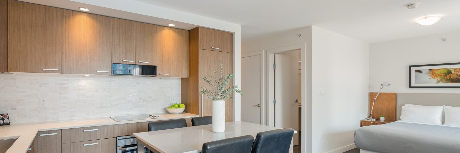 A modern studio apartment featuring a kitchen with wooden cabinets, a white countertop, and a dining table with four chairs. In the background, there’s a bed, a nightstand, and a framed picture on the wall.