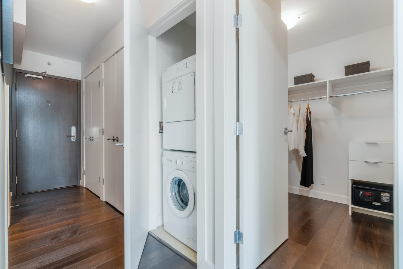 A hallway in a modern apartment features a stacked washer and dryer in a closet, dark wood floors, white walls, and an adjacent walk-in closet with hanging clothes and storage shelves.