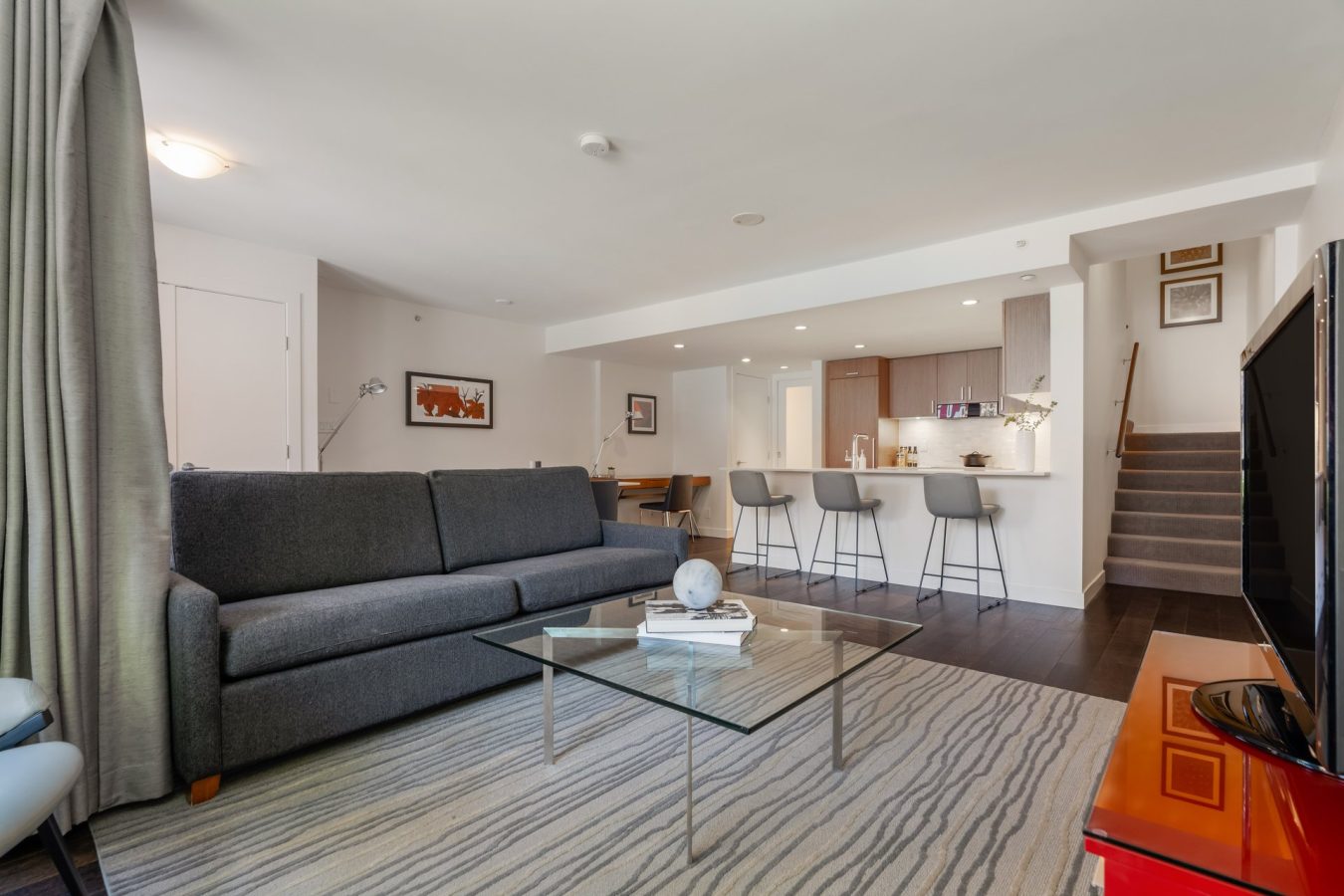 Modern open-plan living room with a dark gray sofa, glass coffee table, TV, striped rug, and kitchen area with bar stools. Stairs and a dining desk are visible; space is bright with neutral colors.